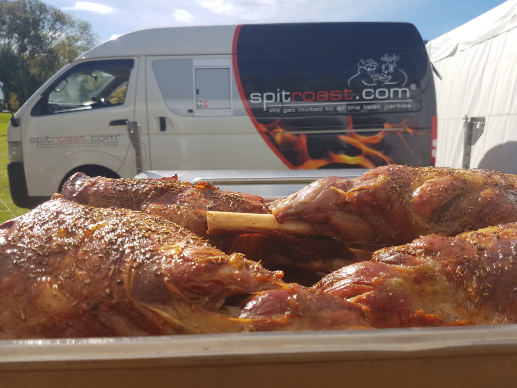 Close up of spitroasted meat on the bone, with a spitroast.com branded van in the background